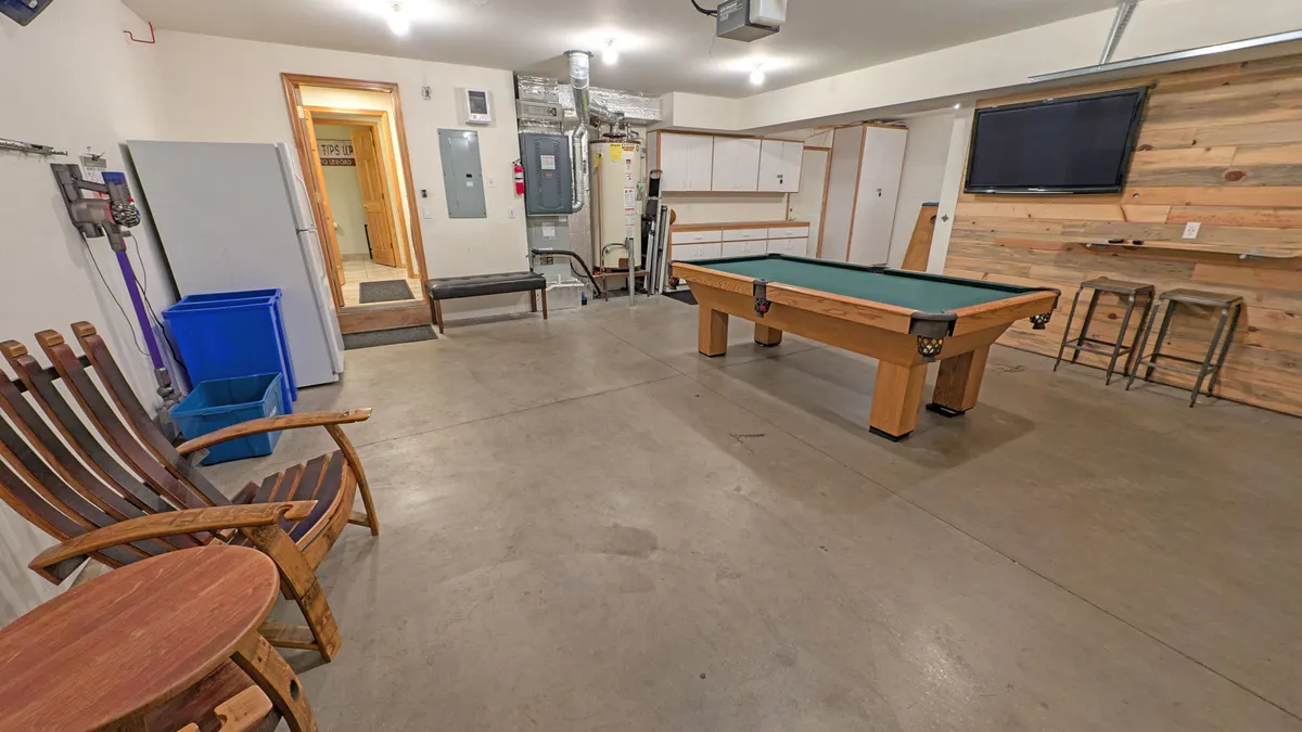 A recreational garage interior with a pool table at the center, wooden chairs and bar stools inviting group gatherings, a wood-paneled wall, and a television mounted for entertainment. The layout encourages social play and active engagement.