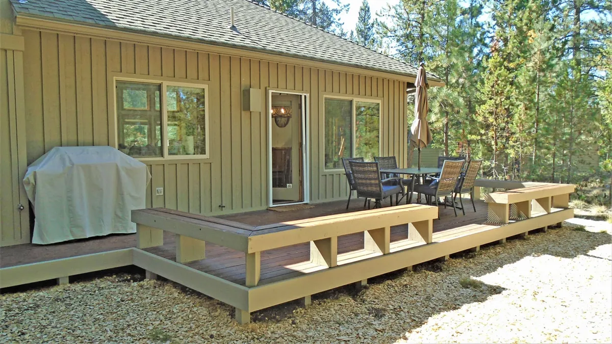 This image shows a spacious deck area with a comfortable outdoor dining set and a grill, set against a tranquil backdrop of trees. The table is shaded by an umbrella, creating a welcoming space for alfresco meals, morning coffees, or evening gatherings. The deck boards and surrounding greenery add to the relaxed, sun-kissed ambiance.