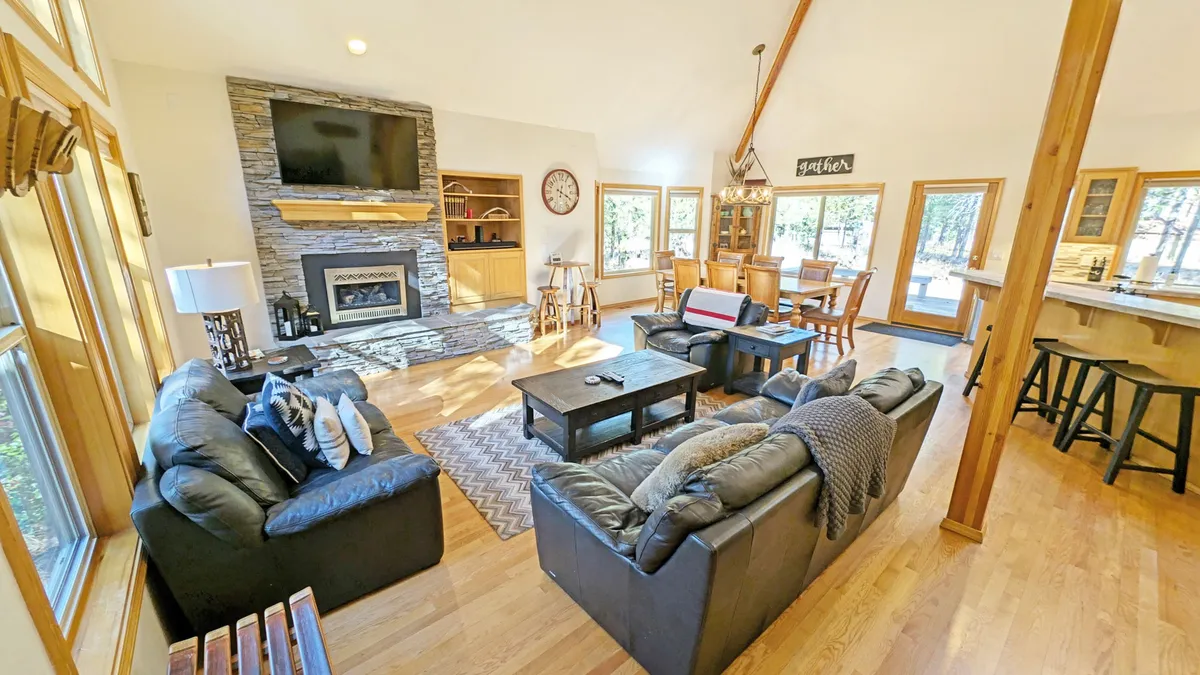 The image showcases a spacious and beautifully appointed living area with a cozy gas fireplace as the centerpiece, plush seating arranged for conversation and relaxation, a generous dining table perfect for group meals, and expansive windows that fill the space with natural light and offer views of the serene outdoors.