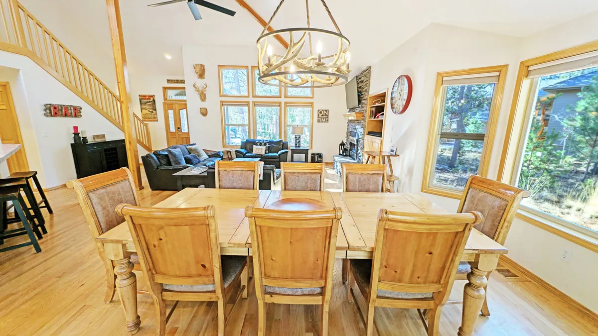 A cozy, well-lit dining and living area with a large dining table ready for family meals or game nights, modern chandelier, ample seating, and windows that bring the outdoors in.