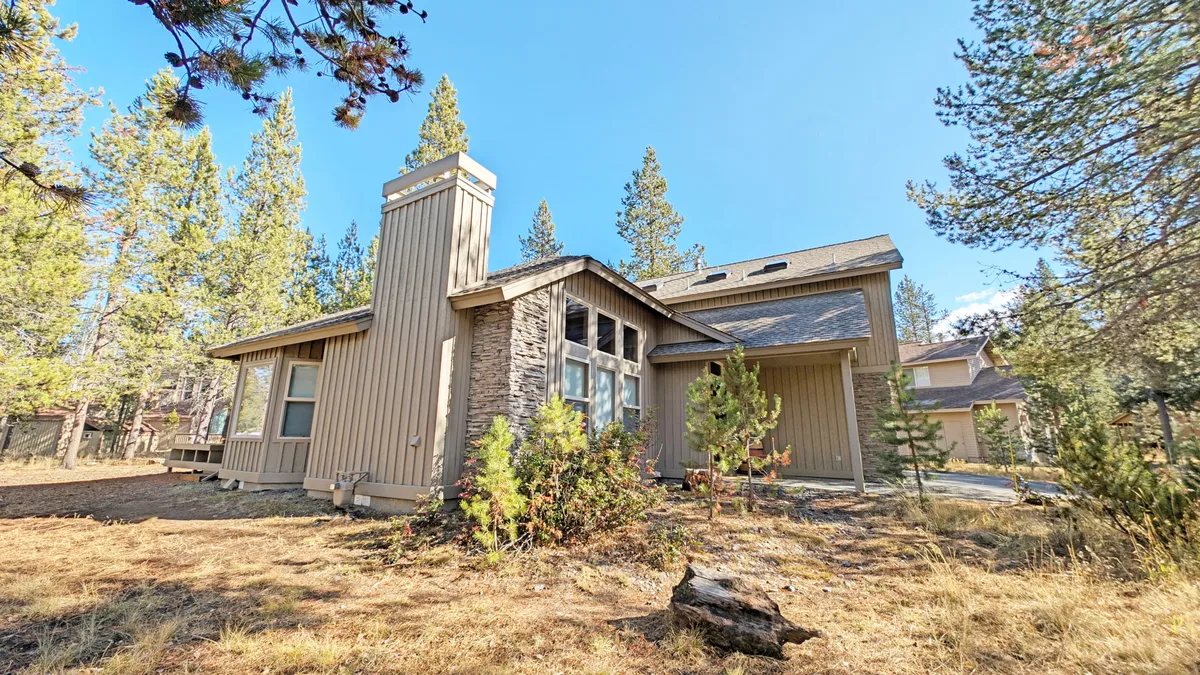 An exterior view of a wooden home with stone accents, large windows, and lush trees all around. The scene is bathed in natural light, exuding tranquility and inviting guests to enjoy the outdoor spaces.