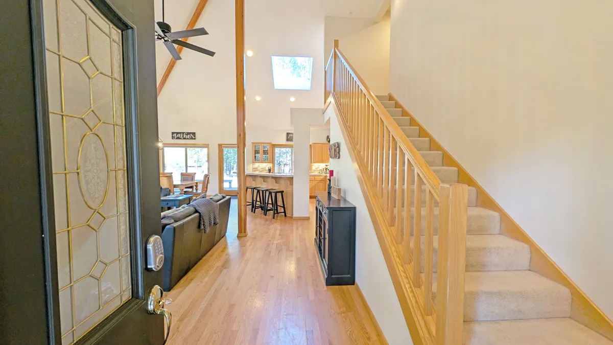 A bright, open-concept living space showing the front door, staircase, kitchen island, and a cozy living area. Large windows fill the room with natural light, inviting guests to unwind, socialize, and enjoy group gatherings.