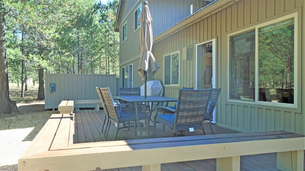 A large outdoor dining table with an umbrella sits on a wooden deck, surrounded by tall trees. The area is set up for family gatherings, barbecues, and lounging, with plenty of space for both socializing and relaxing in the sun or shade.