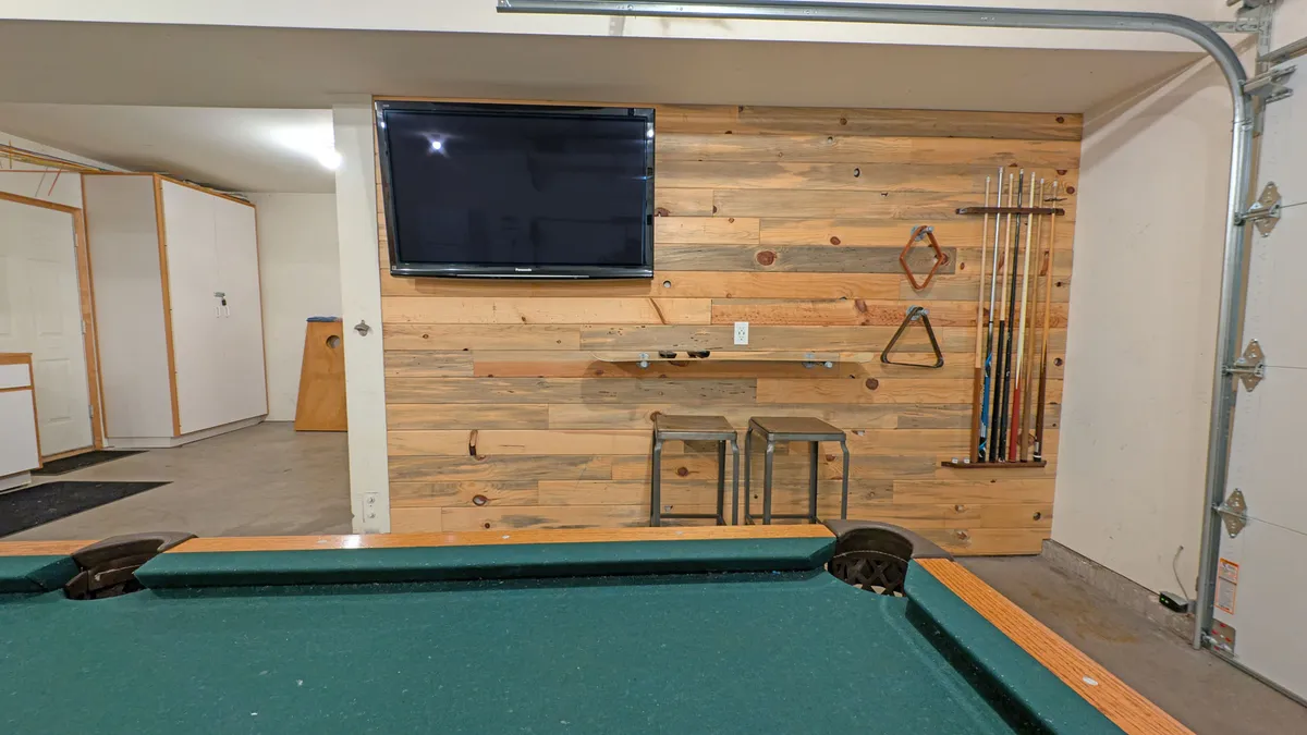 A vibrant garage game room featuring a full-sized pool table, a wall-mounted television, a rustic wooden wall, and bar stools neatly arranged along a built-in bar area. Warm lighting and an inviting atmosphere make this space ideal for both play and socializing.