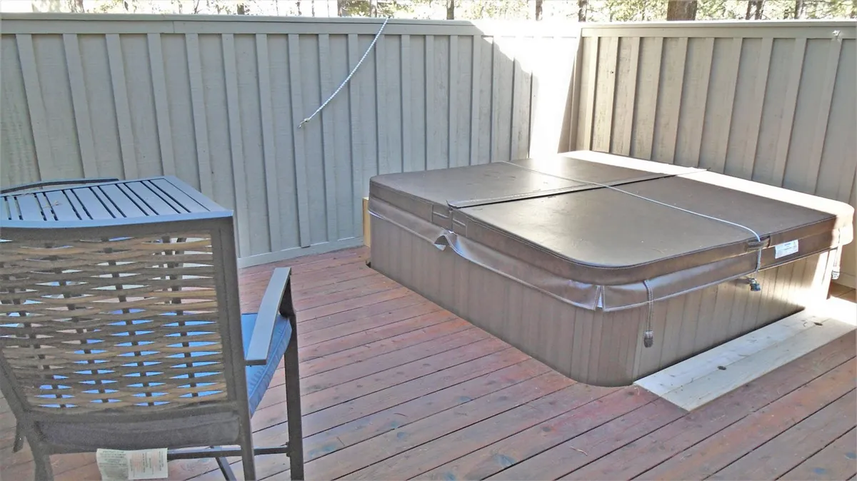 A serene hot tub sits on a wooden deck, with a chair nearby and sunlight filtering through the surrounding trees. The inviting water and forested view suggest a peaceful retreat for soaking, socializing, or stargazing right outside the home.
